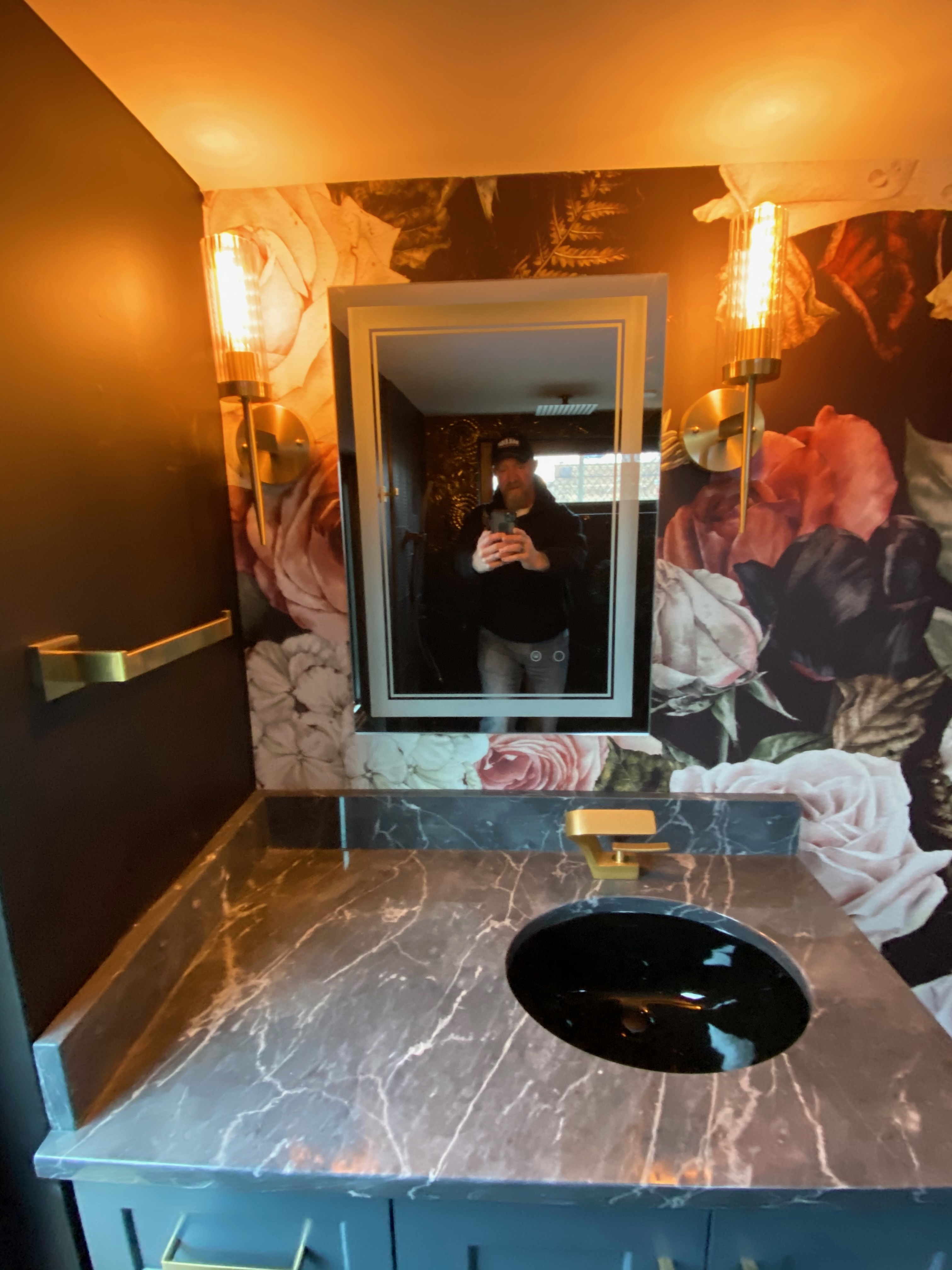 Compact bath vanity with floral wallcovering, dark stone top, and brass lighting
